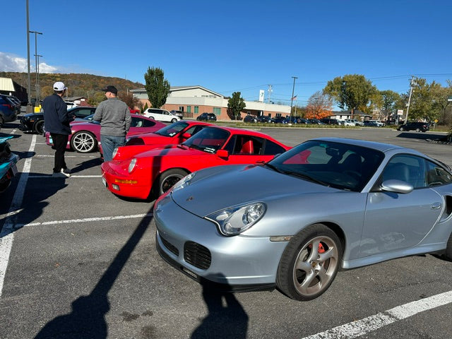 Car club members gathered around classic sports cars in the parking lot, with autumn hills behind
