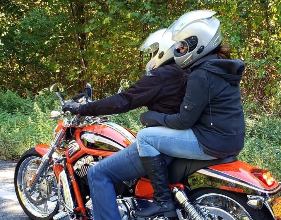 Two riders on an orange and chrome motorcycle on a tree-lined back road